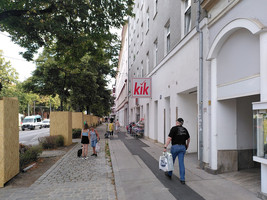 Gehsteig, linke Hälfte gepflastert und etwas tiefer gelegen, rechte Hälfte asphaltiert, auf der linken Hälfte stehen zwei Personen und unterhalten sich, auf der rechten Hälfte gehen Personen mit Einkaufssäcken, vor einem Geschäft stehen Körbe mit Waren