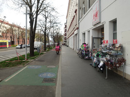 Asphaltierter Radweg und asphaltierter Gehsteig ohne Sitzbänke, vor einem Geschäft stehen Körbe mit Waren