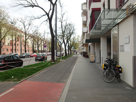 Asphaltierter Radweg und asphaltierter Gehsteig ohne Sitzbänke, links daneben eine Grünfläche und wiederum daneben eine KFZ-Fahrspur, am Gehsteig stehen Fahrräder und ein Schild vor einem Geschäft 